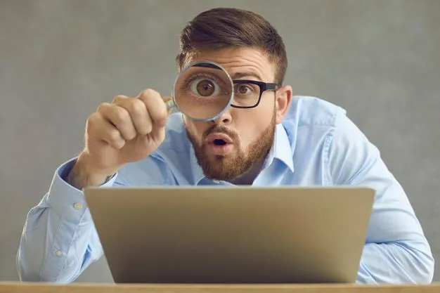 Mann sitzt vor einem Notebook und kontrolliert mit einer Lupe, was er am Bildschirm sieht